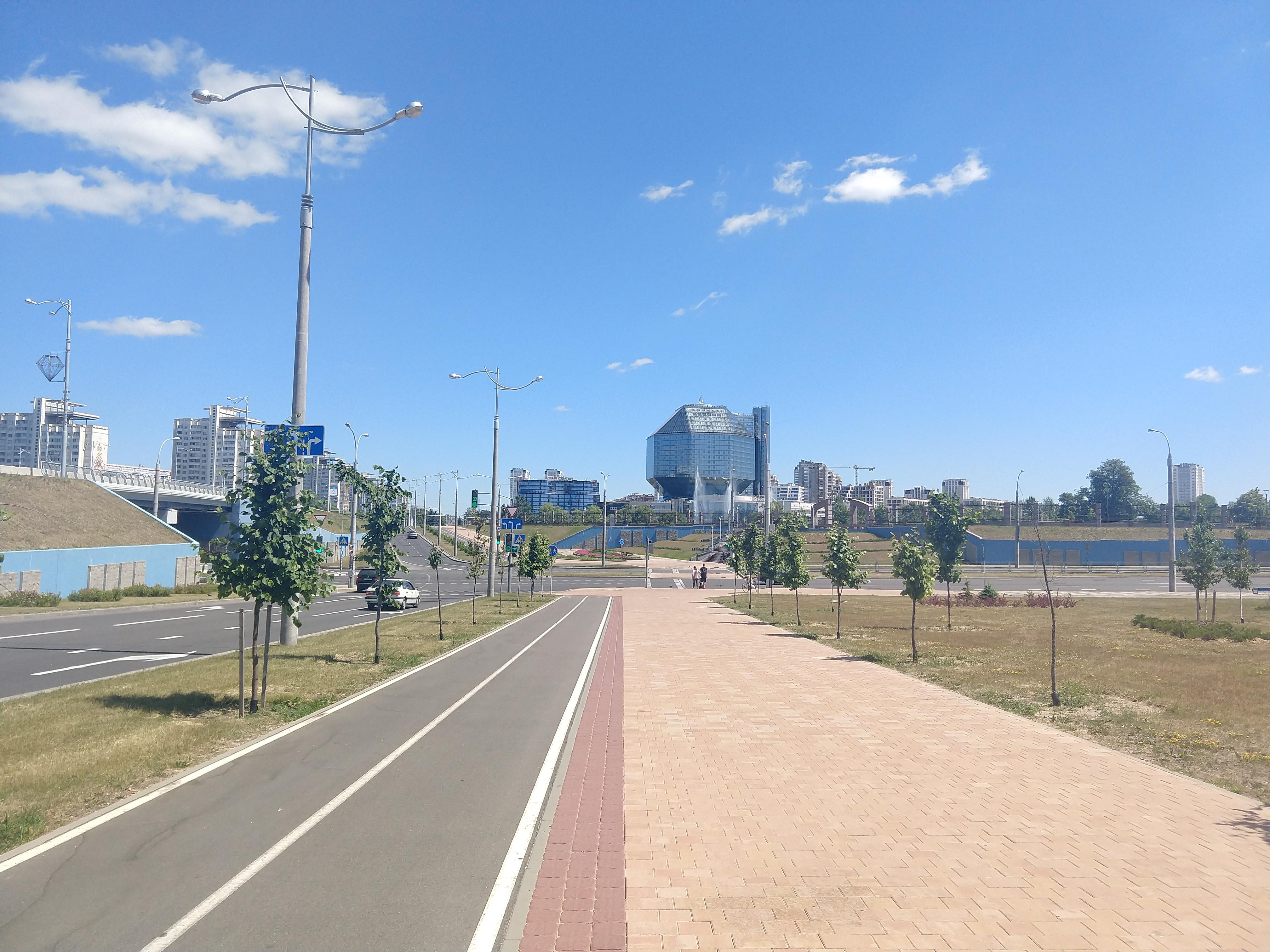 A broad walkway and tree-lined street leading toward a geometric building beneath a blue sky